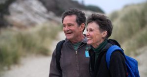 An older couple walking on a beach with a backpack.