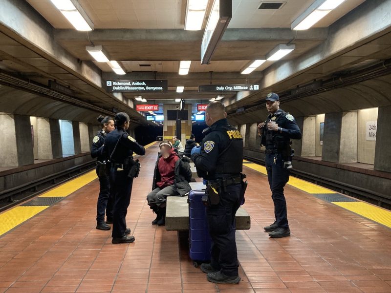 Four police officers standing around a handcuffed woman on a bench in a BART subway station. Someone thought her torch lighter was a gun.