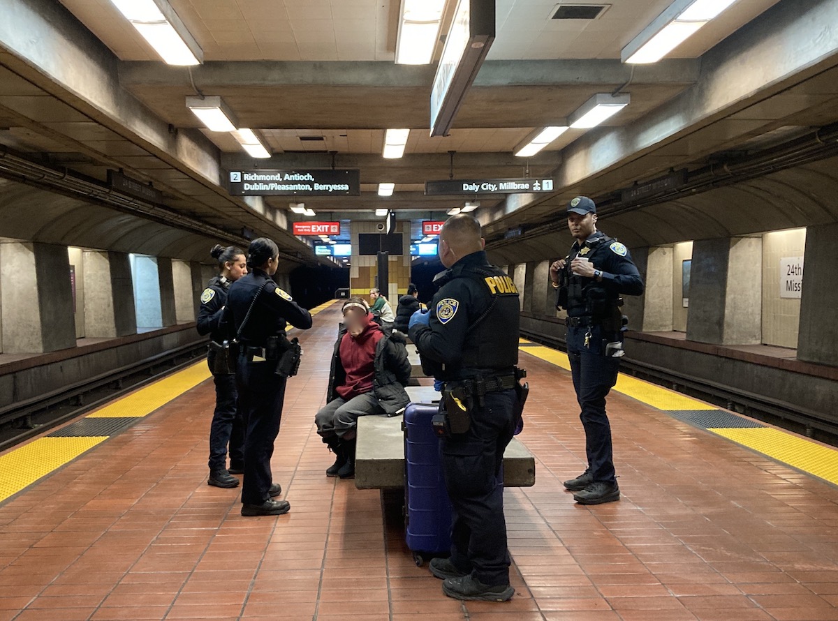 Four police officers standing around a handcuffed woman on a bench in a BART subway station. Someone thought her torch lighter was a gun.