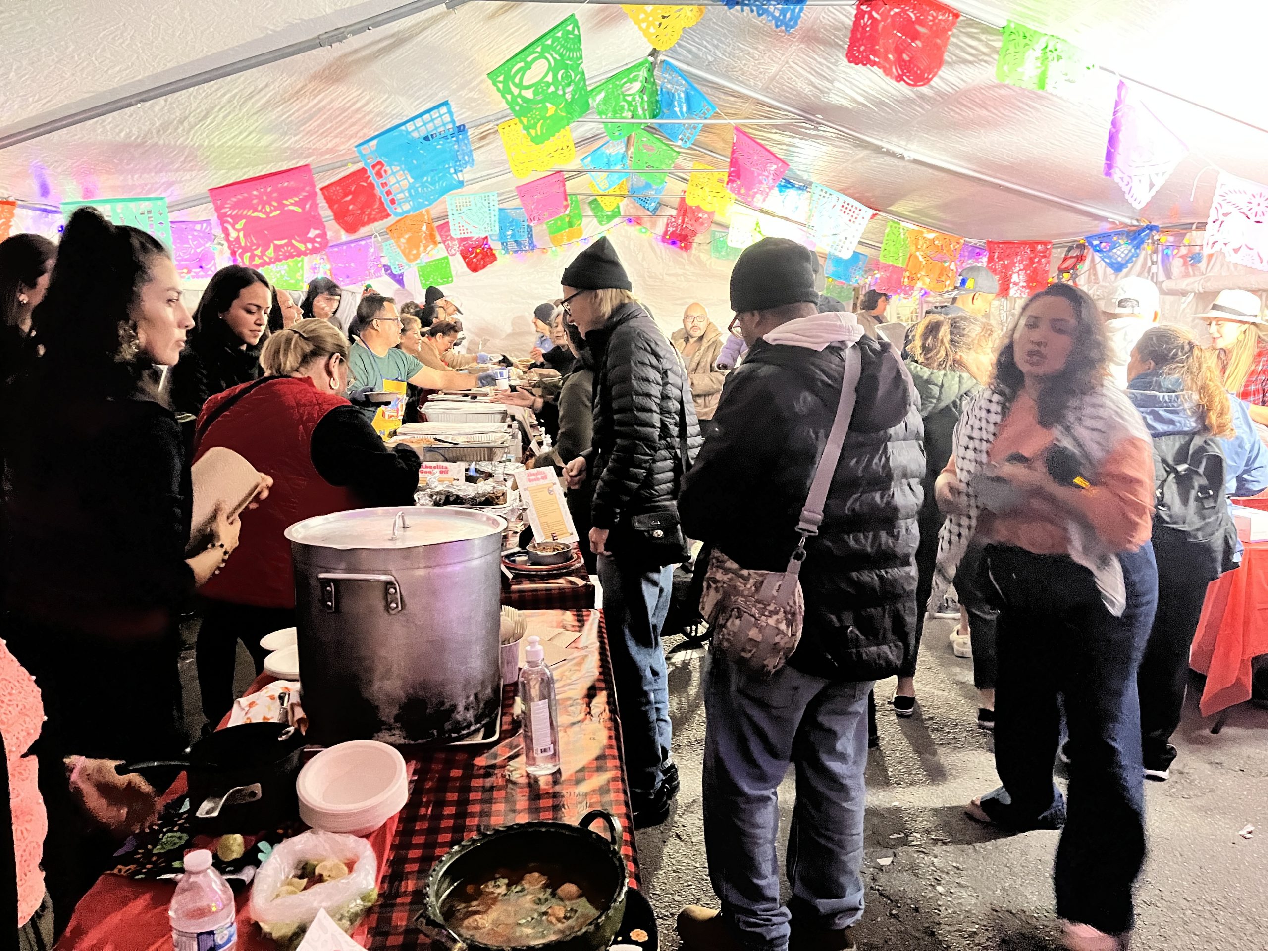 A group of people standing under a tent getting food at the Abuelita Cookoff.