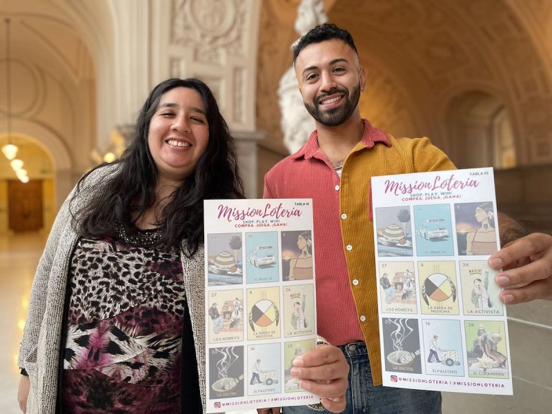 Two people standing in a building holding up mission loteria game boards.
