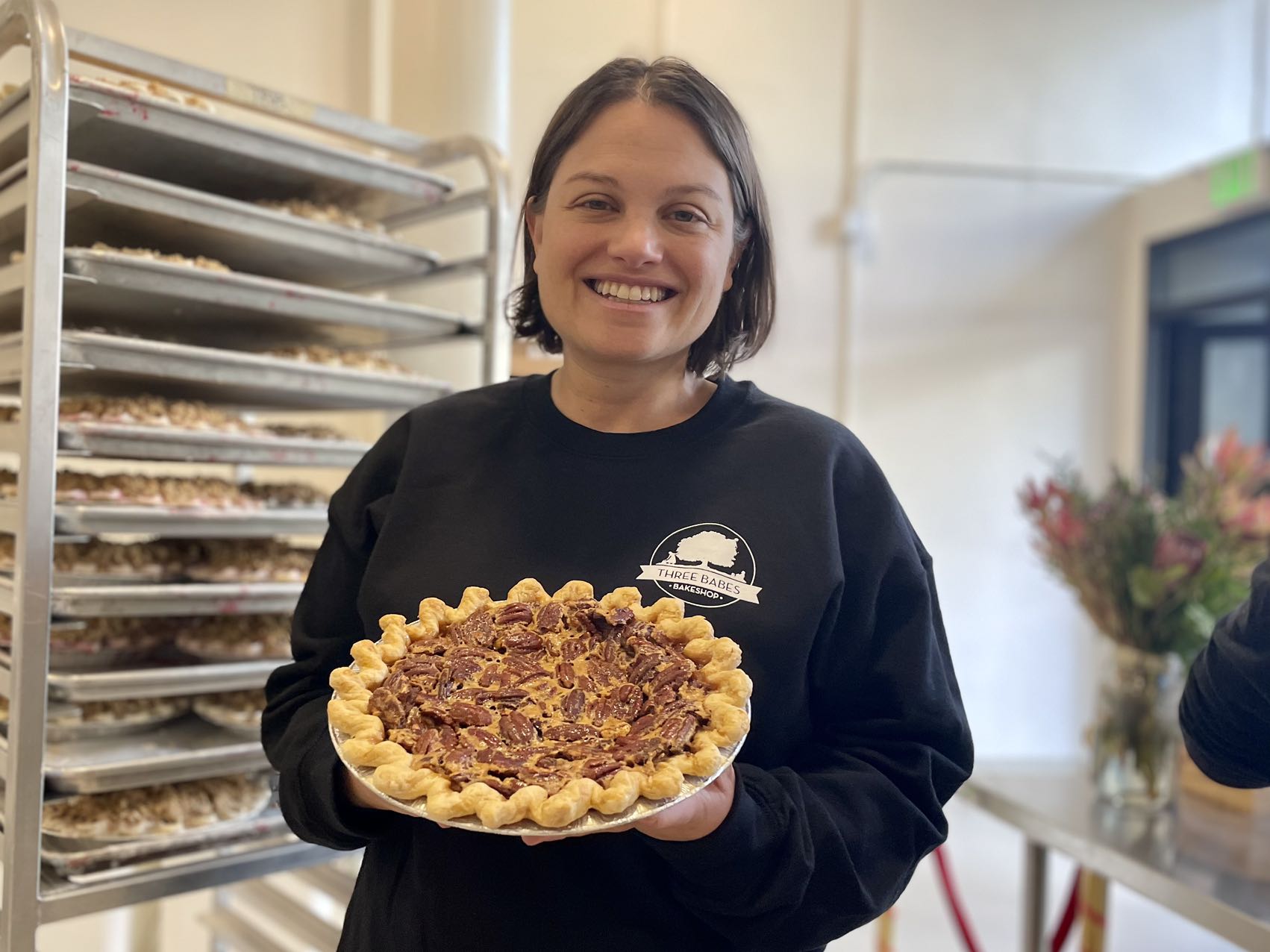 Lenore Estrada holding a pie in front of the Three Babes Bakeshop rack.