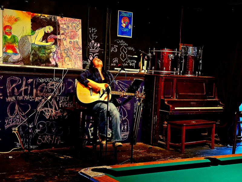 A singer playing an acoustic guitar in front of a pool table on the stage of a bar.
