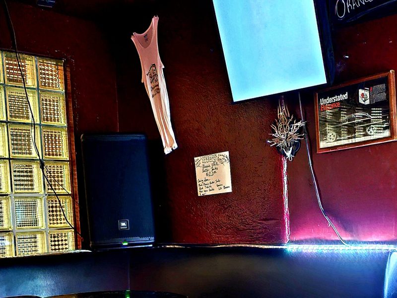 A red wall of a bar with a shirt, some signs and a TV hanging on it. One of the signs says "whiskey 25 cents."
