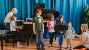 A group of children standing around a piano in a classroom.