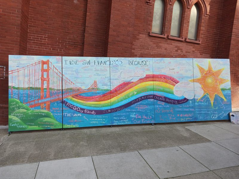 A mural of the Golden Gate Bridge, a rainbow, a cloud, and a sun. "I love San Francisco because ... " is written on the mural and reasons for people to love San Francisco written all over the mural.