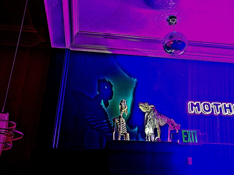 In a room with purple lighting, white neon light sign that says "MOTHER" hangs on the wall.