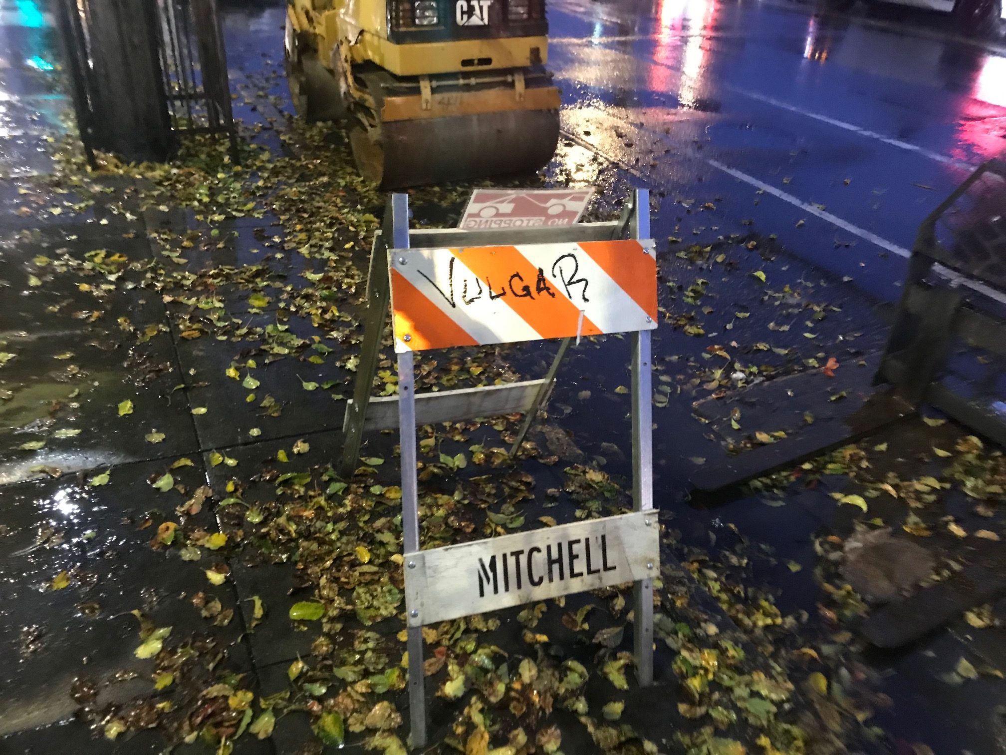 A traffic barricade with the words 'Vulgar' and 'Mitchell' on the wet sidewalk.