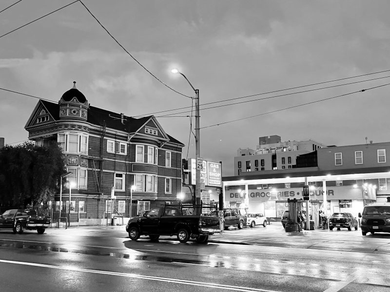A black and white photo of a gas station and a Victorian style house next to it at night.