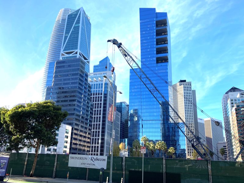 A crane at a construction site in San Francisco. Skyscrapers under the blue sky are in the background.