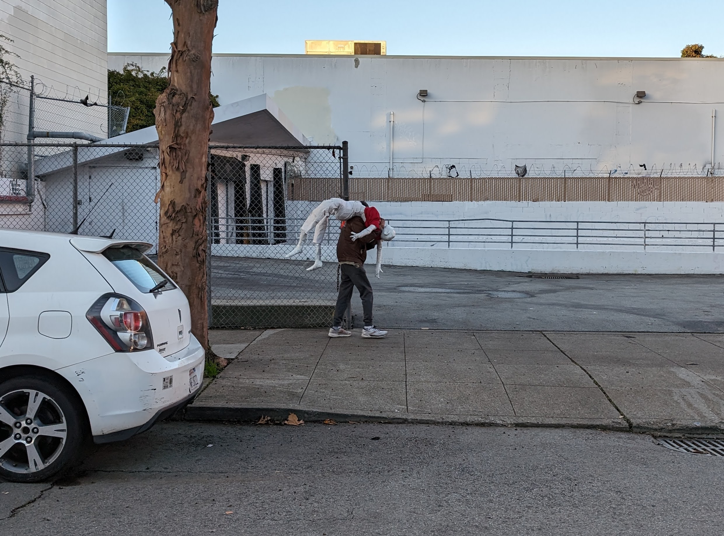 A person walking past a tree carrying a mannequin that is dressed in white pants and a red shirt. A white car parked by the side of the street.