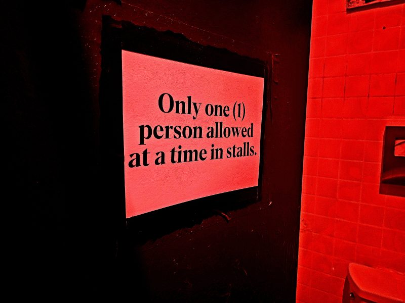 A sign on a wall in the men's bathroom at a bar that says: Only one (1) person allowed at a time in stalls.