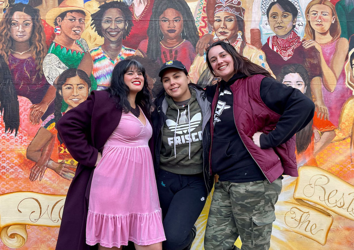 Three women posing in front of a mural of women at Balmy Alley and Lovers Lane.