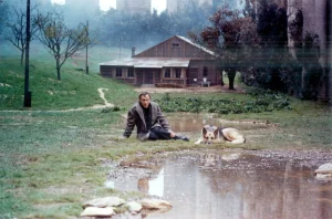 A man sits on the ground next to a puddle with a dog.