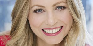 A woman with blond hair and blue eyes is smiling.