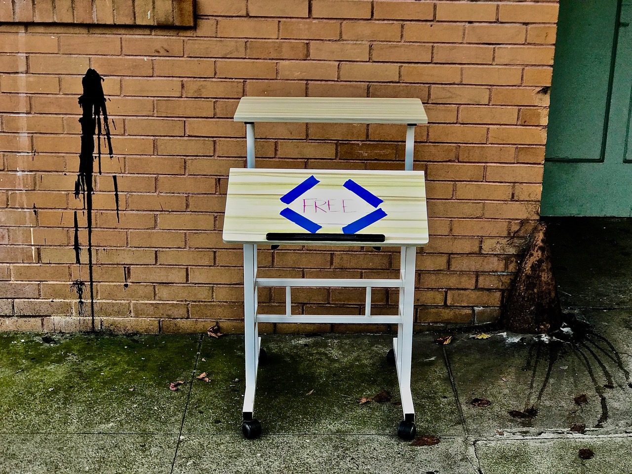 A small desk sitting on a sidewalk in front of a brick building. A sign on it says FREE.