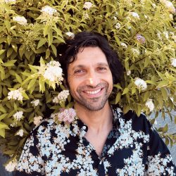 A man in a floral shirt smiling in front of a bush.