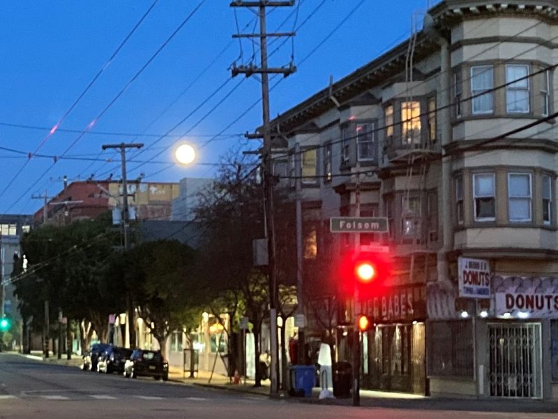 City street at dusk with a full moon rising above, traffic light on red, and illuminated storefront signs.