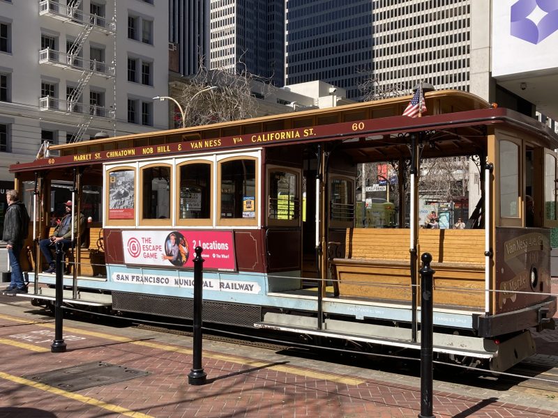 A cable car at a stop in san francisco, with passengers on board and surrounding buildings in the backdrop.