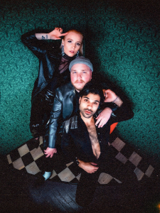 Three individuals posing for a photo with a vintage aesthetic, featuring a patterned backdrop and checkered floor.