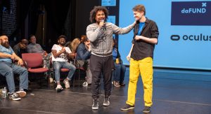 Two men smiling and engaging with each other on stage, one holding a microphone, in front of an audience and a screen with logos.