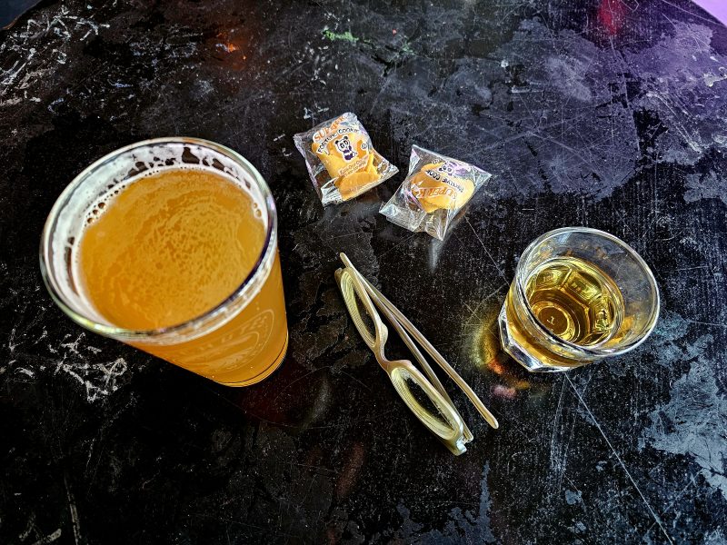 A glass of beer, a shot of whiskey, eyeglasses, and two packaged fortune cookies on a dark tabletop.