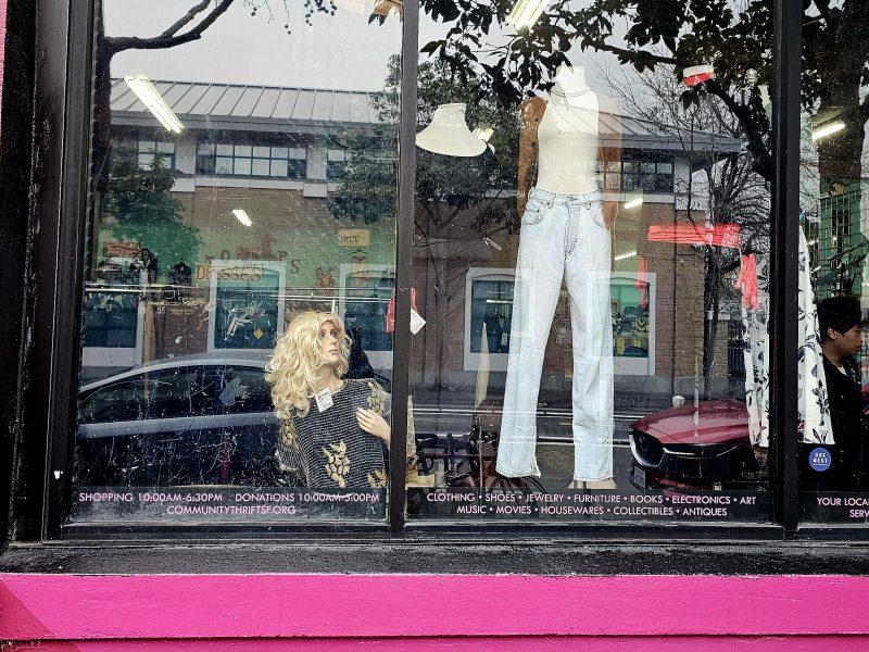 A mannequin with blonde hair wearing a black and white striped top and white pants is displayed in a store window with various text advertisements, while reflections of the street outside are visible on the glass.