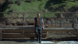 A shirtless man in dark pants and suspenders standing in front of a pen with large animals, likely on a farm.