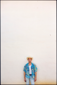 Man in a cowboy hat and tropical shirt standing against a plain background.