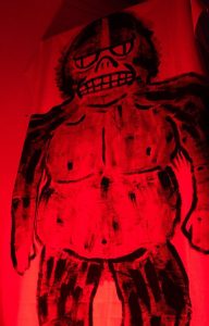 Red-tinted image of a drawing of a humanoid figure with exaggerated features on a wall.