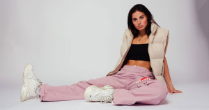 Woman sitting on the floor wearing pink pants, a black top, a beige jacket, and white sneakers.