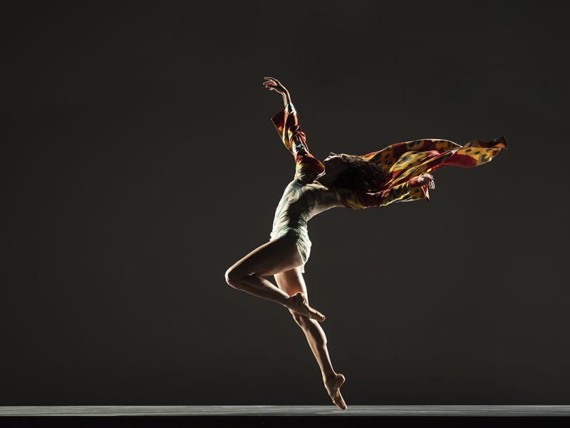 Alonzo King LINES Ballet's Ilaria Guerra captured mid-twirl, with a colorful shawl flowing behind her against a dark background.