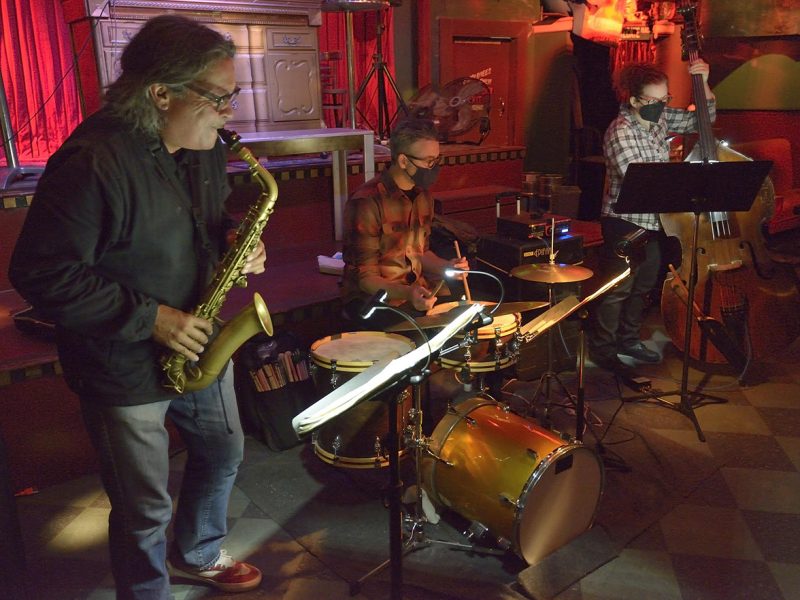 A saxophonist and a drummer performing on stage with a seated guitarist and another musician with sheet music in the background.