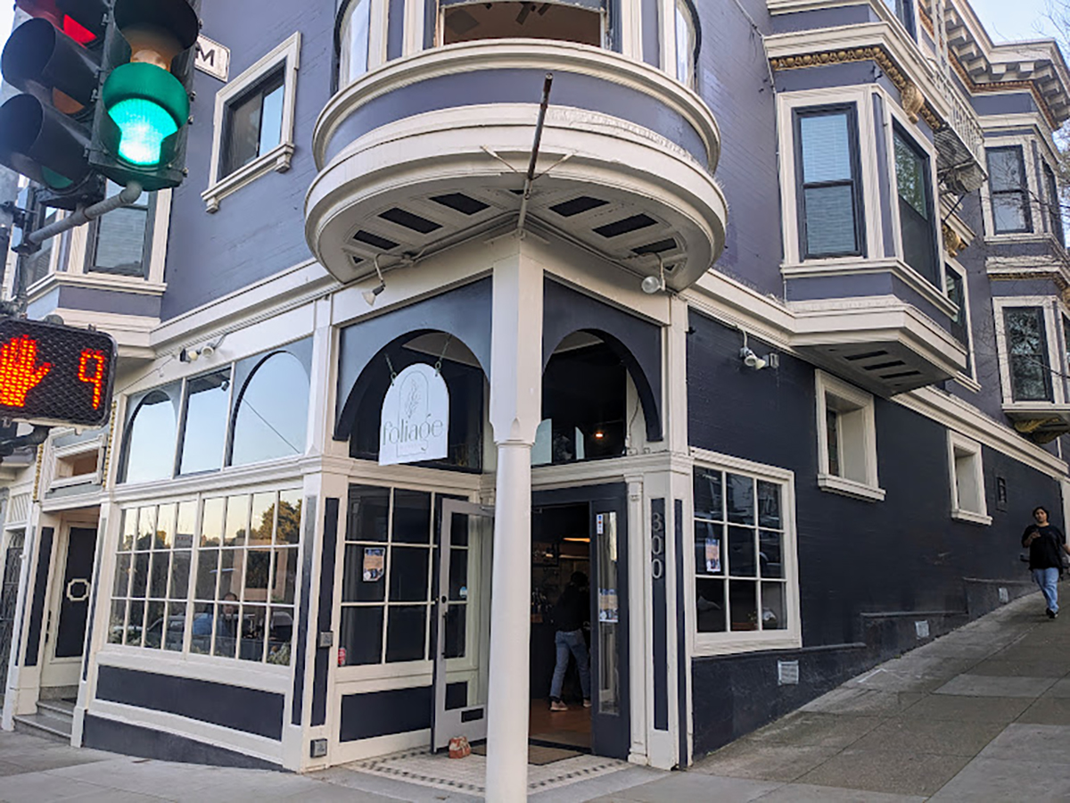 A curved corner building with white and navy blue exterior, hosting a business named 'melanie's,' with a pedestrian walking by and a traffic light showing green.