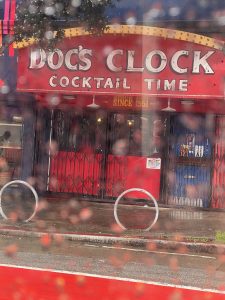 Rain droplets on a window with a view of doc's clock bar, which advertises "cocktail time" and notes its establishment since 1951.