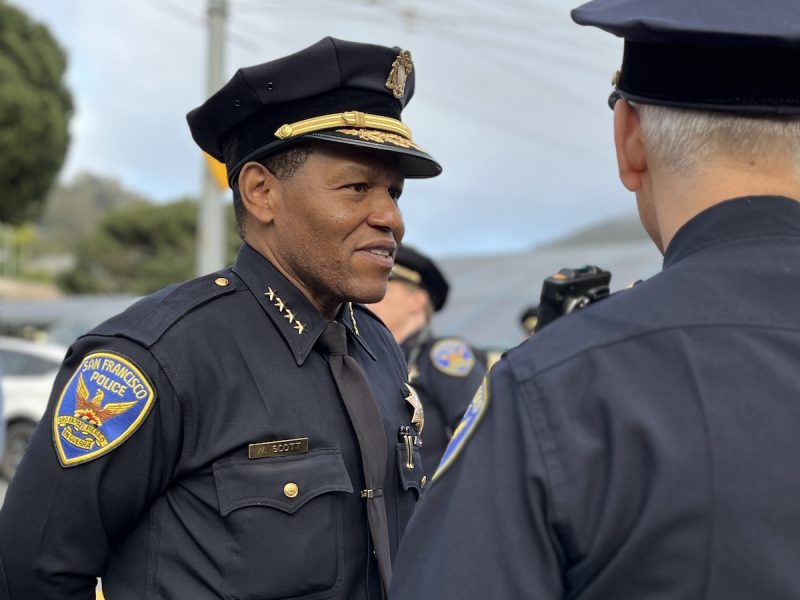 A uniformed police officer with captain's bars on the collar conversing with a colleague outdoors.