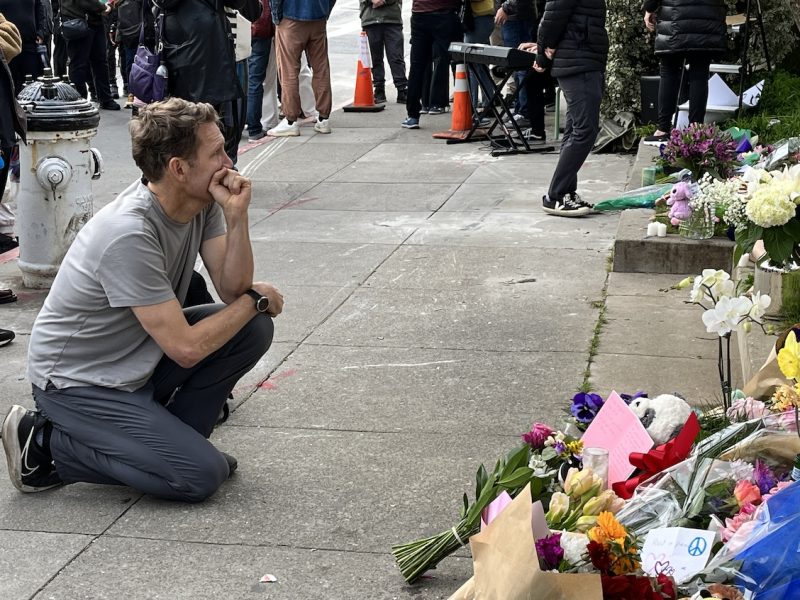 A person crouching and looking thoughtful in front of a makeshift memorial with flowers.
