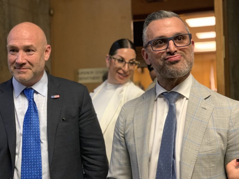 Three individuals posing in a hallway, with the man on the right looking up and to the side, the woman in the center slightly out of focus and smiling, and the man on the left looking directly at the camera.