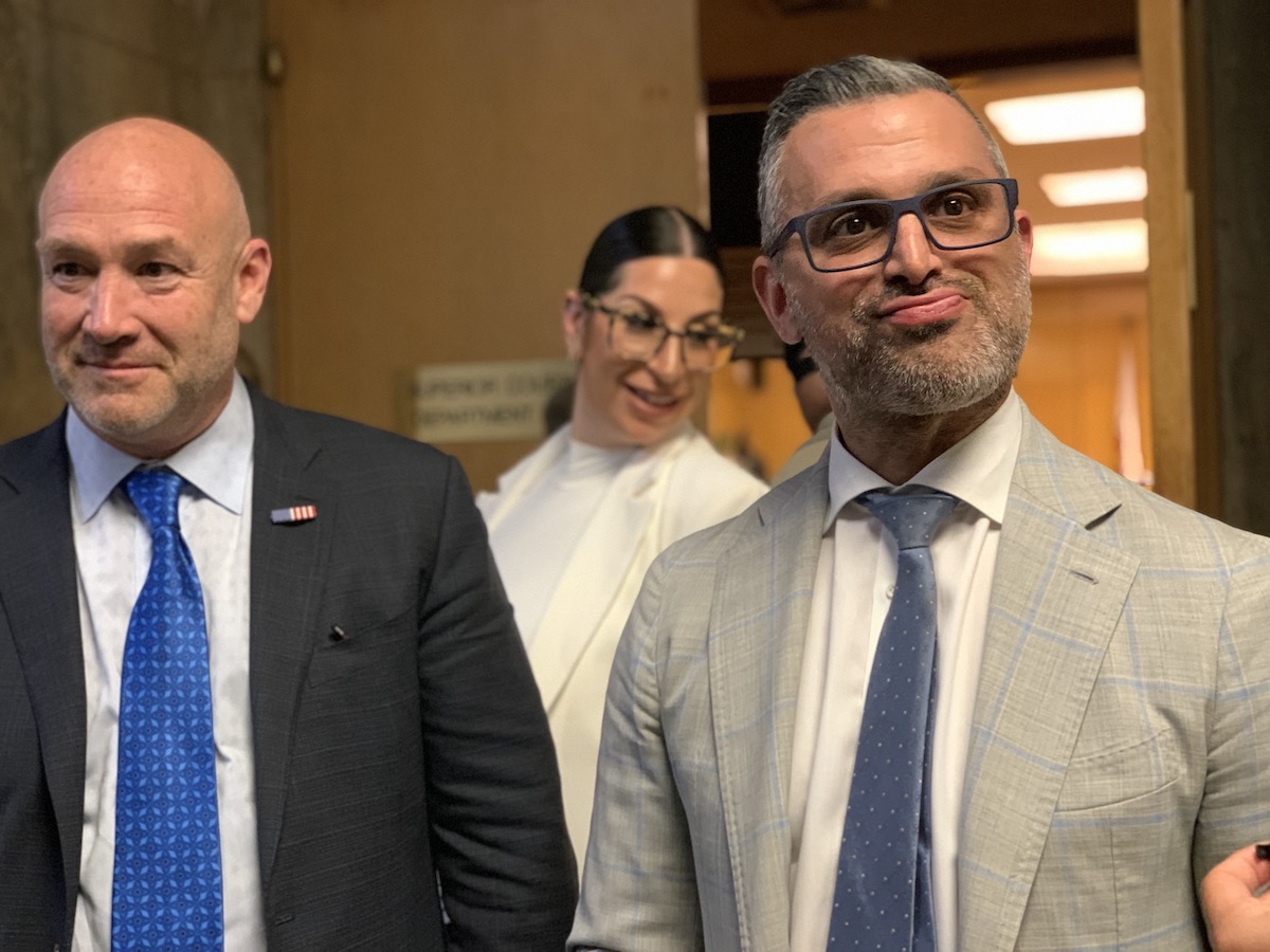 Three individuals posing in a hallway, with the man on the right looking up and to the side, the woman in the center slightly out of focus and smiling, and the man on the left looking directly at the camera.