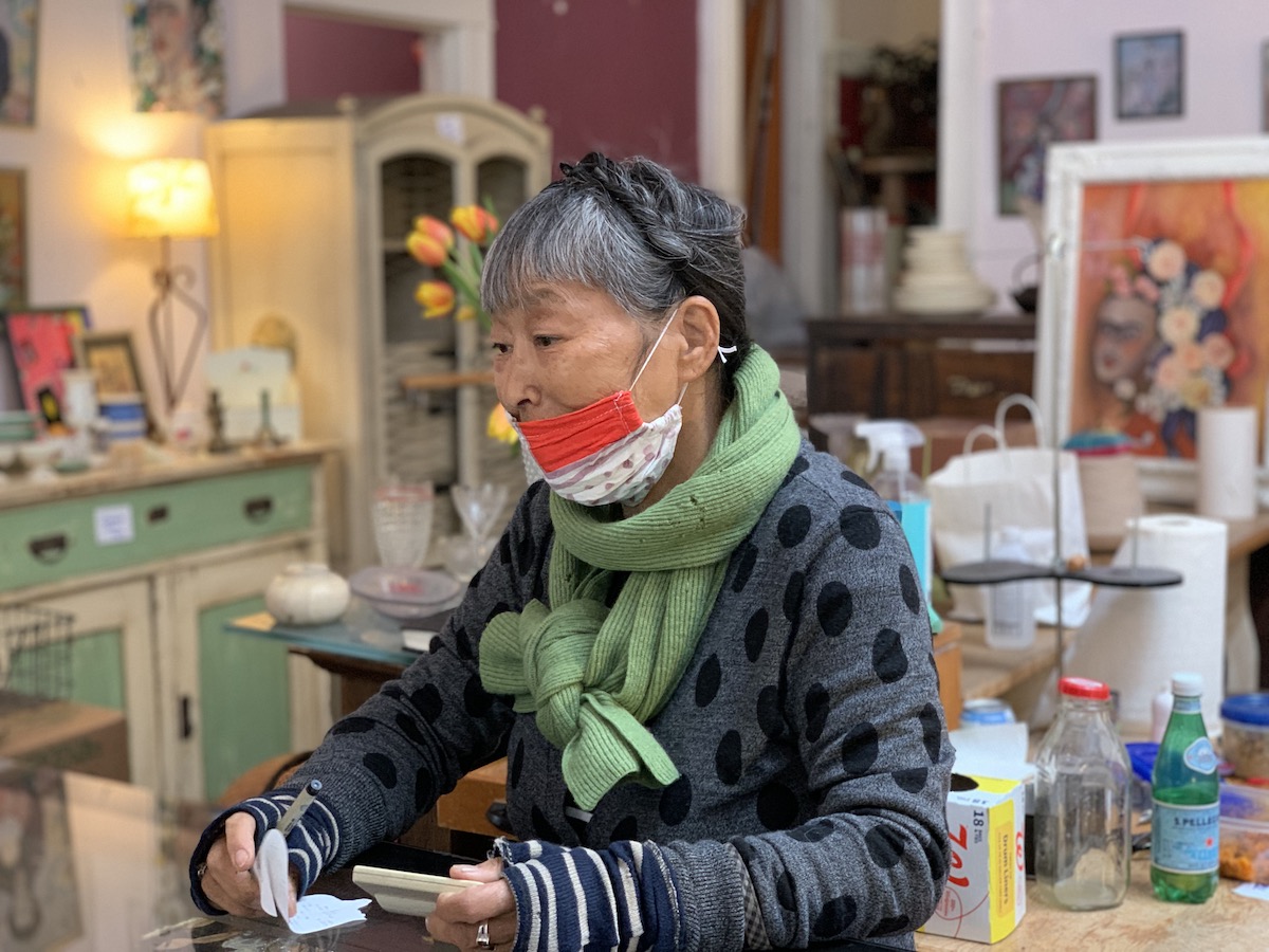An older woman with a face mask around her chin writing on a piece of paper in a room with colorful furniture and paintings.