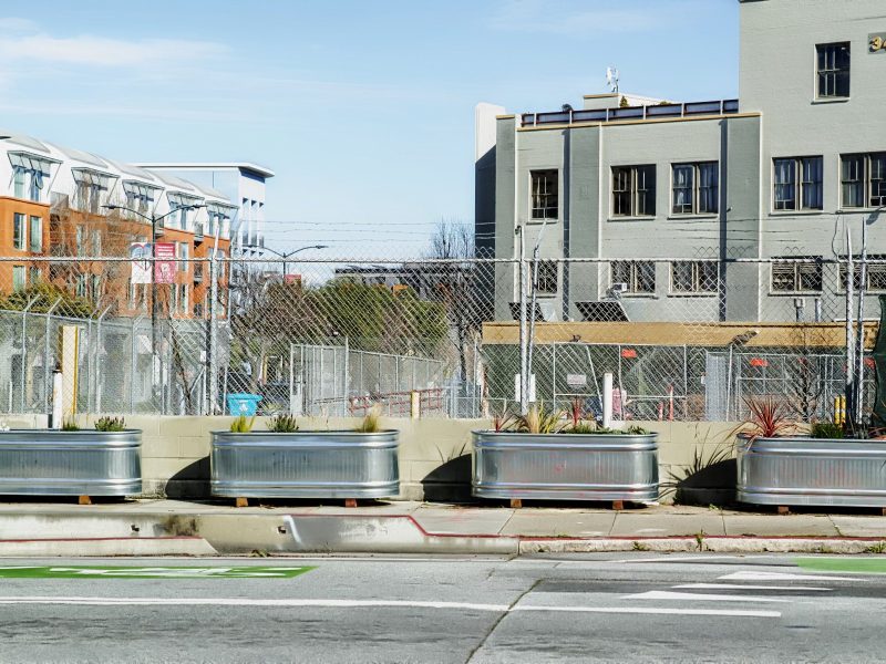 Urban street with large planters, chain-link fence, and mixed-use buildings in the background.