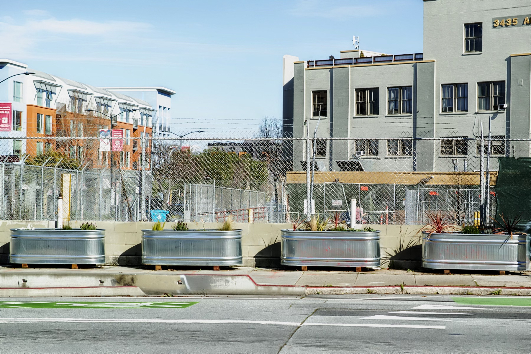 Urban street with large planters, chain-link fence, and mixed-use buildings in the background.