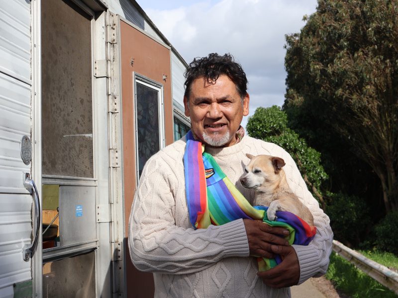 A man wearing a cream sweater and a colorful scarf stands outdoors holding a small dog wrapped in a matching scarf, with a trailer in the background.