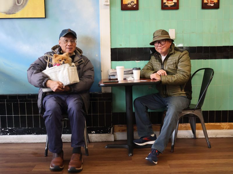 Two individuals sitting at separate tables in a Claddagh-themed cafe, with one holding a small dog in a carry bag.