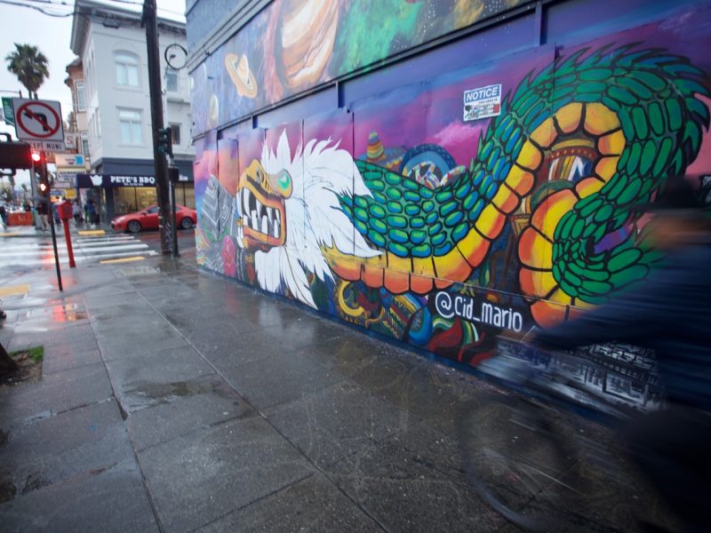 A colorful mural of a dragon on an urban street wall with a pedestrian passing by in motion blur.