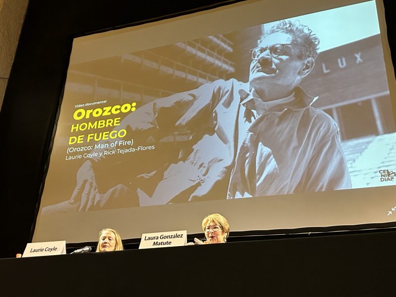 Two speakers at a panel discussion on the film "orozco: hombre de fuego" pictured on a screen above.