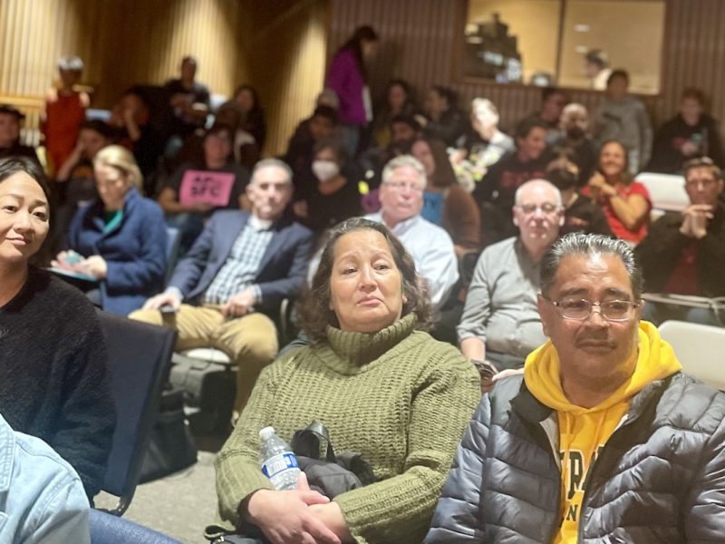 A group of people seated in a hall facing the camera, some looking attentively forward, with a person holding a sign in the background.