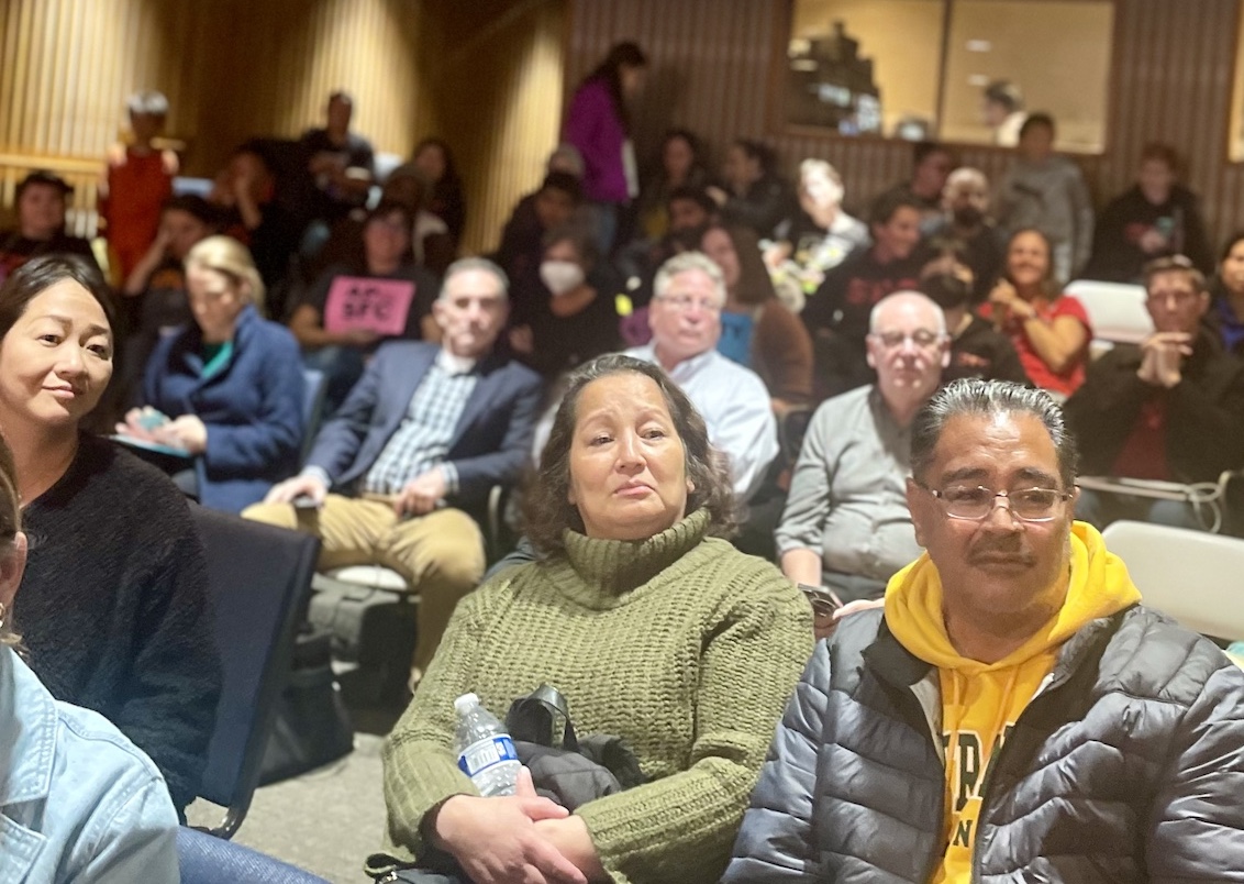 A group of people seated in a hall facing the camera, some looking attentively forward, with a person holding a sign in the background.