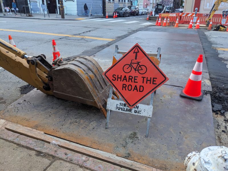 Construction zone with a "share the road" sign next to a backhoe bucket and traffic cones.