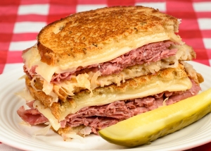 A reuben sandwich with corned beef, sauerkraut, and melted cheese, served with a pickle on the side.